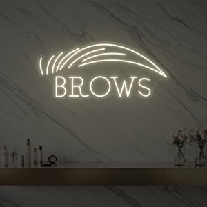 Könnte beinhalten: Ein weißes Neon-Schild in Form einer Augenbraue mit dem Wort „Brows“. Das Schild ist an einer Marmorwand befestigt. Darunter befindet sich ein goldener Tresen mit Schönheitsprodukten und Blumen.