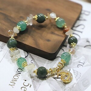 May include: A green and gold beaded bracelet with a crescent moon charm. The bracelet features various green and white stones, as well as a gold charm with a crescent moon and a small white stone.
