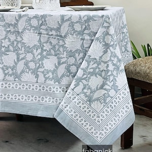 May include: A light blue tablecloth with a white floral pattern covers a wooden table. The tablecloth has a border with a geometric design. A water pitcher and glasses are on the table.