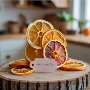 Puede incluir: Rodajas de naranja y naranja sanguina secas dispuestas sobre una rodaja de madera. Una pequeña etiqueta con el texto "cherie emilia" se coloca frente a la fruta. Las rodajas varían en color de amarillo claro a rojo intenso.
