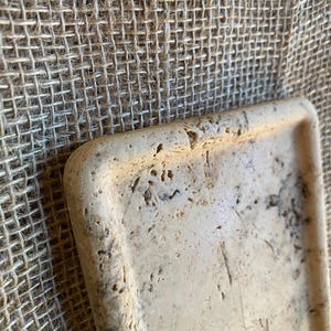 May include: A rectangular tray made of light beige travertine stone with a decorative design on the bottom. The tray has a rough, textured surface.