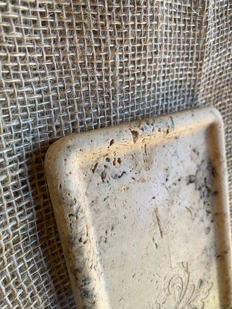 May include: A rectangular tray made of light beige travertine stone with a decorative design on the bottom. The tray has a rough, textured surface.