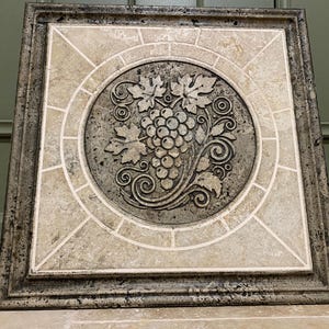 May include: A square tile with a decorative design of a grapevine and grapes in a circle. The tile is made of beige and brown stone with a textured surface.