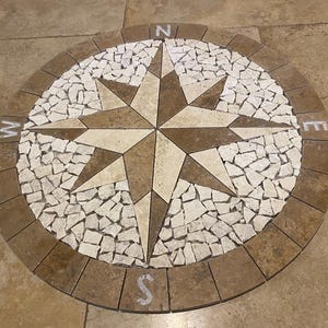 Peut inclure: Une rose des vents circulaire en mosaïque dans les tons marron, beige et blanc. Les points cardinaux sont marqués des lettres N, E, S et W. La mosaïque est faite de carreaux de pierre et est encastrée dans un sol en pierre.