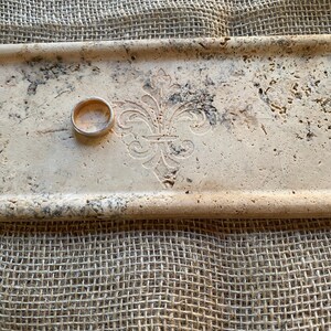 May include: A rectangular tray made of light beige travertine stone with a decorative fleur-de-lis design. A gold ring is resting on the tray.
