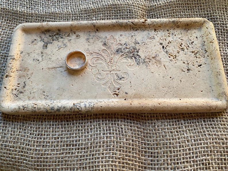 May include: A rectangular tray made of light beige travertine stone with a decorative fleur-de-lis design. A gold ring is resting on the tray.