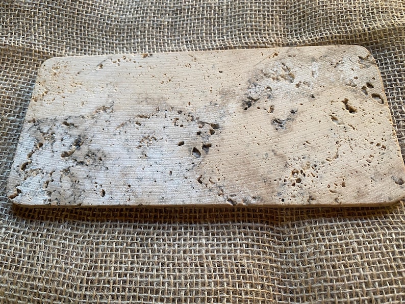 May include: A rectangular, light beige travertine tile with a rough, textured surface. The tile has a natural, organic look with a variety of small holes and crevices.