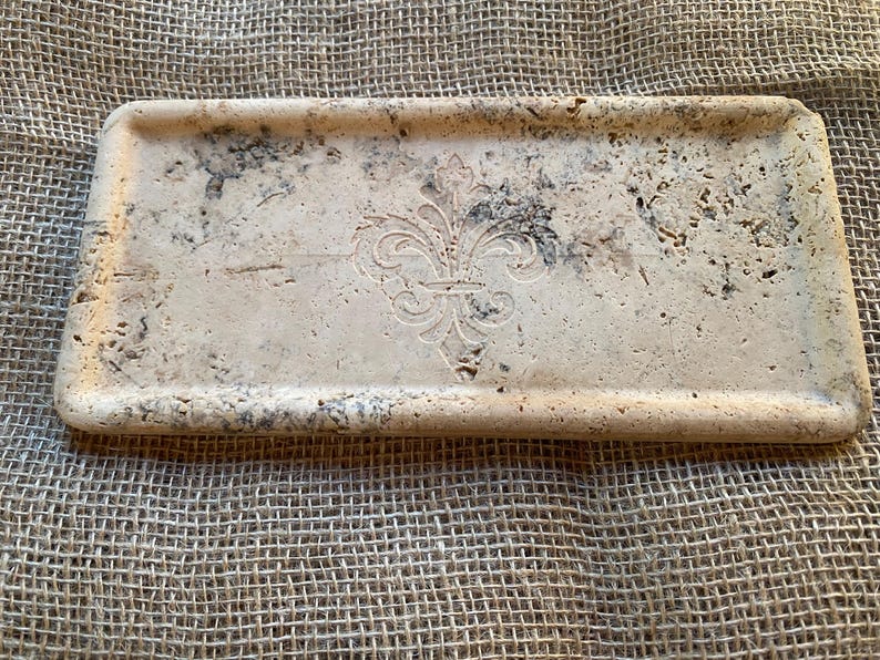 May include: A rectangular tray made of light beige travertine stone with a raised, carved fleur-de-lis design in the center.