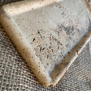 May include: A rectangular tray made of light beige travertine stone with a raised, decorative design in the center. The tray has a slightly rough texture and a beveled edge.