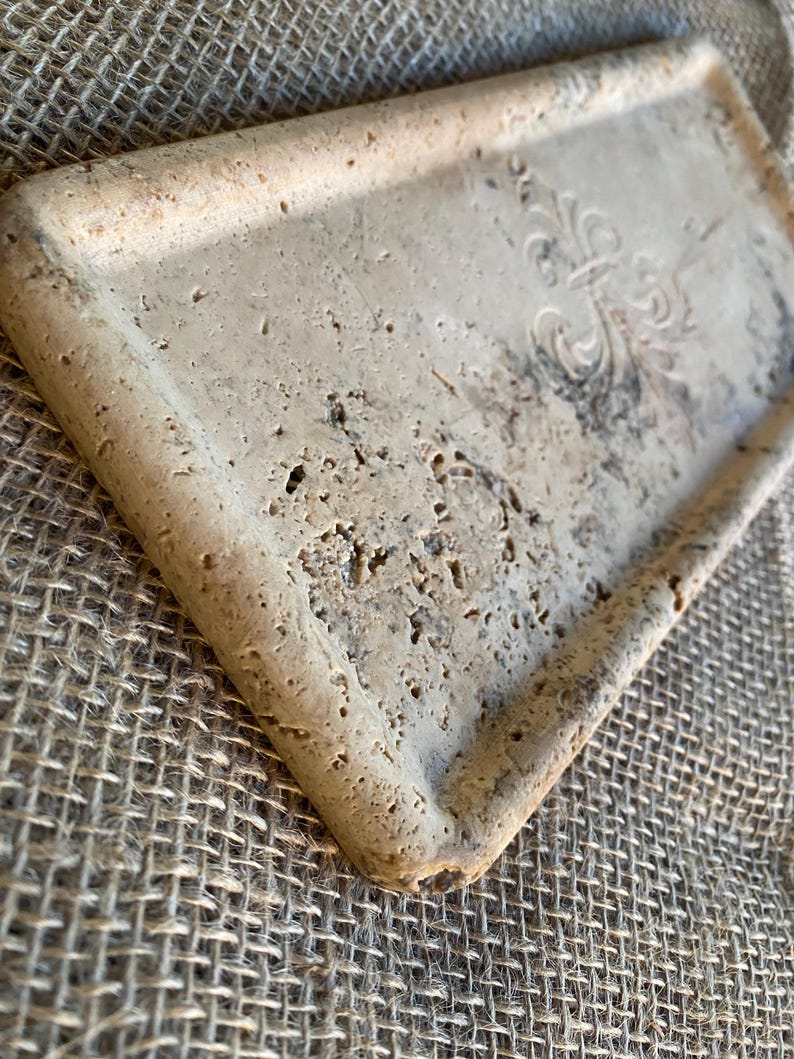May include: A rectangular tray made of light beige travertine stone with a raised, decorative design in the center. The tray has a slightly rough texture and a beveled edge.