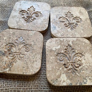 Puede incluir: Juego de cuatro posavasos cuadrados de piedra beige con un diseño de flor de lis.