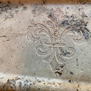 May include: A close-up of a light beige stone tray with a carved fleur-de-lis design.