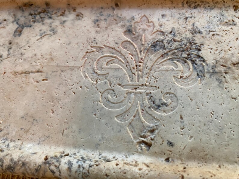 May include: A close-up of a light beige stone tray with a carved fleur-de-lis design.