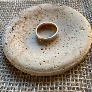 May include: A silver ring with a thin band and a wider band on the outside, resting on a beige stone coaster.