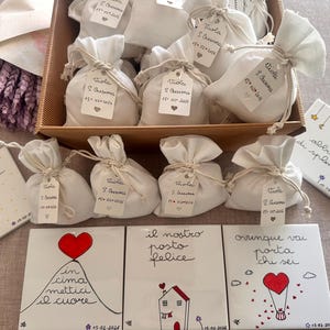 Può includere: Una collezione di piccoli sacchetti di stoffa bianca legati con spago, ognuno con un'etichetta. Le etichette hanno testo scritto a mano e un cuore. Sono presenti anche diverse piastrelle di ceramica decorate con testo scritto a mano e illustrazioni.