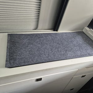 Könnte beinhalten: Eine rechteckige, graue Filzmatte mit schwarzem Rand liegt auf einer weißen Oberfläche. Die Matte besteht aus einem gesprenkelten, strukturierten Material. Das Bild ist aus leicht erhöhter Perspektive aufgenommen.
