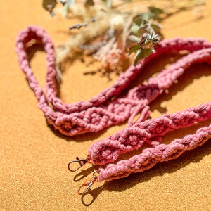 Könnte beinhalten: Ein staubrosa Makramee-Lanyard mit zwei bronzefarbenen Drehverschlüssen. Das Lanyard besteht aus dickem, gewebtem Baumwollkordel. Der Hintergrund ist eine strukturierte, hellbraune Oberfläche mit getrockneten Blumen.