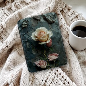 May include: A tablet case with a floral design, featuring a large, open rose and smaller buds in pink and cream. The case has a textured, dark grey background and is on a beige knitted blanket. A white mug of coffee is also visible.
