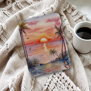 May include: A rectangular case featuring a sunset scene with palm trees, ocean, and a vibrant sky in shades of orange, pink, and yellow. The case is resting on a beige knitted blanket, with a white mug of coffee in the background.