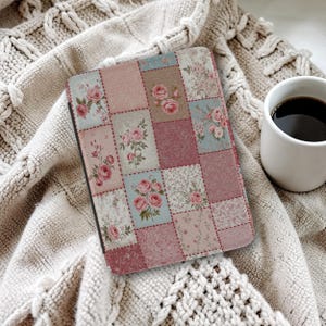 May include: A tablet case with a patchwork design, featuring squares of pink, blue, and beige with rose patterns. It rests on a cream knitted blanket. A white mug of coffee is in the background.