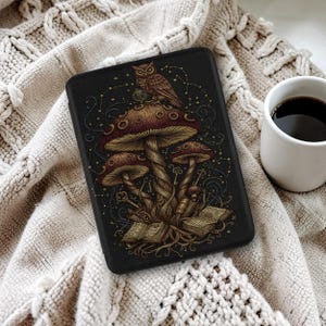 May include: Black tablet case featuring a whimsical design of mushrooms, an owl, keys, and books. The artwork is in shades of brown, gold, and blue, set against a dark background. A white mug of coffee sits to the right, on a cream-colored knitted blanket.