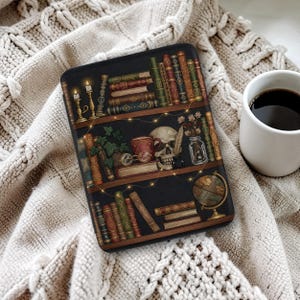 May include: A black tablet case with a detailed illustration of a bookshelf with antique books, candles, a skull, and a globe. The case is on a cream knitted blanket, with a white mug of coffee in the background.