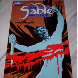Puede incluir: Portada de cómic que presenta una figura con piel azul, sosteniendo un cuchillo, con un diseño rojo similar a la sangre en el pecho. El título "Jon Sable Freelance" se muestra en una fuente estilizada, con el logo de First Comics en la parte superior.