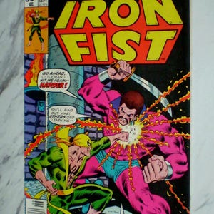 Puede incluir: Portada de cómic de Iron Fist con una escena de acción dinámica. El título "Iron Fist" está en amarillo con un contorno rojo. Dos figuras luchan, una en verde y amarillo, la otra en rosa y azul. El cómic está fechado en septiembre.