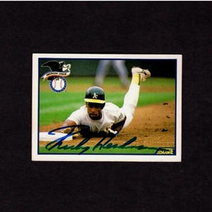 May include: Baseball card showing a player sliding into a base. The card includes the American League logo and a signature. The player is in a white and green uniform and a black helmet.