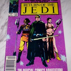 May include: A comic book cover for Star Wars: Return of the Jedi, issue #1. The cover features three characters in the center, with a purple background and the title in yellow. The Marvel logo and issue details are in the upper left corner. The text "THE OFFICIAL COMICS ADAPTATION!" is at the bottom.
