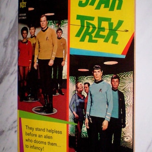 May include: A vintage Star Trek comic book cover. The cover features the Star Trek logo in green on a yellow background. The image shows the Star Trek crew in their uniforms. Text on the cover reads "They stand helpless before an alien who dooms them... to infancy!"