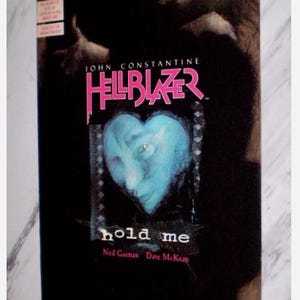Op de afbeelding: Stripboekcover voor "Hellblazer" met een blauw hartvormig gezicht en de woorden "hold me". De titel is roze, met het DC Comics-logo linksboven. De achtergrond is donker met een wazige afbeelding.