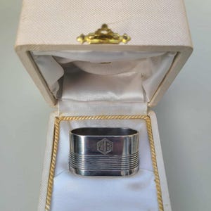 Può includere: Un portatovagliolo d'argento con un design geometrico e iniziali incise, presentato in una scatola color crema. La scatola ha una chiusura dorata e il testo "CHRISTOFLE PARIS" è visibile.