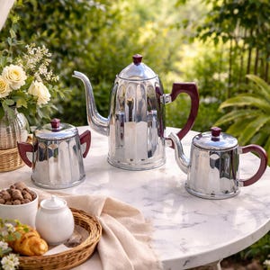 Könnte beinhalten: Ein Set aus drei polierten silbernen Tee- oder Kaffeekannen mit weinroten Griffen und Knäufen. Die Kannen stehen auf einem weißen Marmortisch mit einem Weidenkorb mit Gebäck und einer Blumenvase im Freien.
