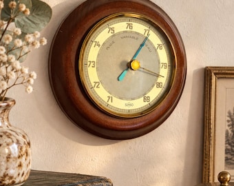 May include: A circular wall-mounted barometer with a dark wood frame and a brass-toned face. The dial features black numerical markings from 69 to 79, the text "VARIABLE", "PLUIE", and "BEAU", and a central yellow cap with two blue hands.