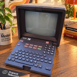 Könnte beinhalten: Ein dunkelgraues Minitel 1 Terminal mit integriertem Bildschirm und AZERTY-Tastaturlayout. Das Gerät verfügt über einen Ziffernblock, Funktionstasten mit orangefarbenen und grünen Akzenten sowie den Schriftzug Minitel 1 auf der Vorderseite.