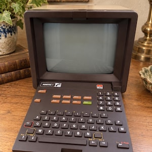 Peut inclure: Un terminal Minitel 1 vintage de marque Alcatel avec un boîtier en plastique marron foncé, un écran intégré et un clavier AZERTY doté de touches orange, vertes et noires. L'appareil comprend un pavé numérique et divers boutons de fonction.