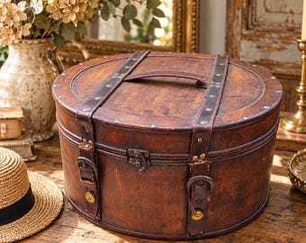 May include: A round, dark brown wooden storage box with a weathered finish. The lid features a central handle and two vertical leather straps secured with metal rivets and buckles. A small metal latch is positioned at the front center of the box.