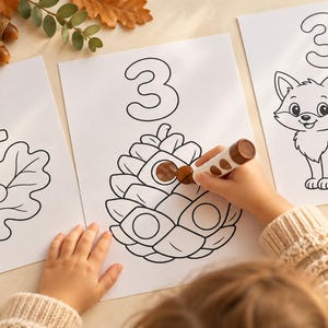 May include: Three white sheets of paper featuring black line art for colouring. The first shows the number 1 and an oak leaf, the second shows the number 3 and a pinecone, and the third shows the number 3 and a fox. A brown marker is colouring the pinecone.