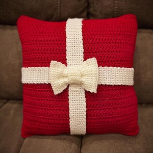 May include: Red crocheted cushion with a white crocheted bow and ribbon, resembling a wrapped gift.