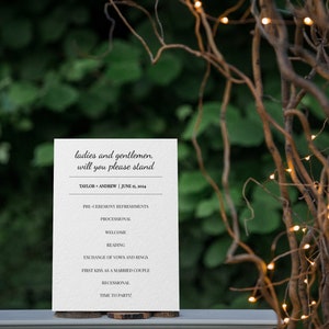 May include: A white wedding program with black text, including the names Taylor and Andrew, and the date June 15, 2024. The program is displayed on a wooden base, with a blurred green background and decorative lights.