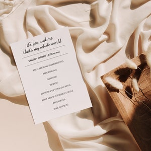 May include: A wedding program with the text "it's you and me, that's my whole world" is displayed on a cream-colored fabric. The program lists the ceremony's order, including "Processional" and "Exchange of Vows." A wooden board with dried flowers rests nearby.