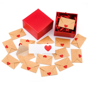 May include: A red gift box, open red box, and several small brown paper envelopes with red heart stickers. A white card with a red heart is also present. The items are arranged on a white surface, creating a romantic theme.