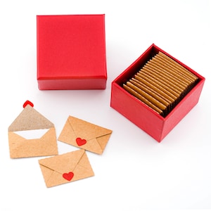 K&ouml;nnte beinhalten: Eine rote quadratische Geschenkbox mit Deckel und eine passende offene Box, gef&uuml;llt mit kleinen, rechteckigen Karten. Drei kleine, braune Papierumschl&auml;ge mit roten Herzsiegeln sind in der N&auml;he verstreut. Ein Umschlag ist ge&ouml;ffnet.