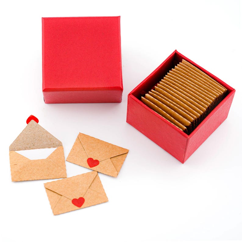 K&ouml;nnte beinhalten: Eine rote quadratische Geschenkbox mit Deckel und eine passende offene Box, gef&uuml;llt mit kleinen, rechteckigen Karten. Drei kleine, braune Papierumschl&auml;ge mit roten Herzsiegeln sind in der N&auml;he verstreut. Ein Umschlag ist ge&ouml;ffnet.
