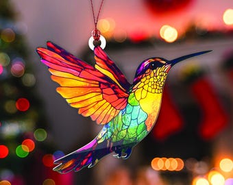 May include: A multi-coloured hummingbird decoration, featuring shades of red, orange, yellow, green, and blue. The bird hangs from a red string and a white ring. The wings are outstretched, and the background is blurred with festive lights, creating a bokeh effect.