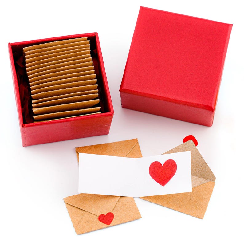 K&ouml;nnte beinhalten: Eine rote Geschenkbox mit einem Stapel kleiner, rechteckiger, brauner Karten. Daneben eine geschlossene rote Box und mehrere kleine braune Umschl&auml;ge und eine wei&szlig;e Karte, jeweils mit einem roten Herzaufkleber.