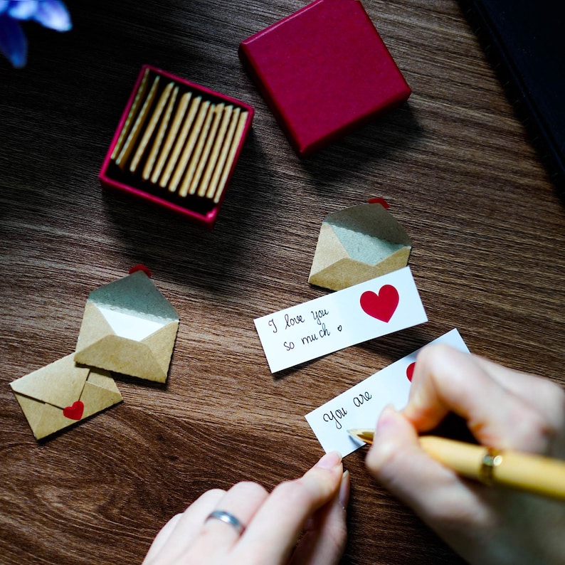 K&ouml;nnte beinhalten: Romantische Szene mit Miniatur-Papierumschl&auml;gen mit roten Herzakzenten, zusammen mit handgeschriebenen Notizen mit den Worten "I love you so much" und "You are". Eine rote Schachtel und eine Streichholzschachtel sind ebenfalls vorhanden.