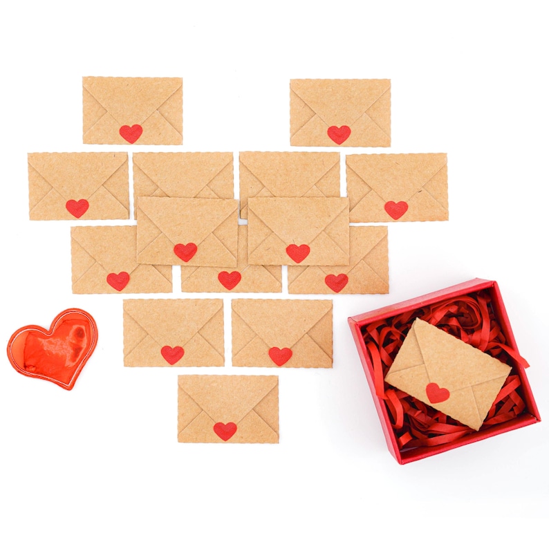 K&ouml;nnte beinhalten: Eine Anordnung kleiner, brauner Papierumschl&auml;ge in Herzform, jeder mit einem roten Herzaufkleber. Rechts befindet sich eine rote Geschenkbox mit rotem Papier und einem Umschlag, links eine herzf&ouml;rmige rote Dekoration.
