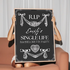 Könnte beinhalten: Ein schwarzes Schild mit Holzrahmen, weißen dekorativen Elementen und dem Text "R.I.P. Emili's SINGLE LIFE BACHELORETTE PARTY." Das Schild wird vor einer Person gehalten, die auf einem pfirsichfarbenen Sofa sitzt.